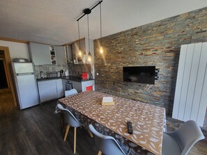 A modern kitchen with a built-in oven, a dining table with chairs, and a stone wall with a TV. - Apartment with balcony and mountain view (Montgenèvre)