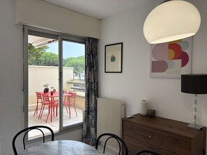 Una habitación con una mesa de comedor, sillas, un radiador, un armario, una lámpara y vistas a un espacio exterior con sillas rojas y una planta en maceta.