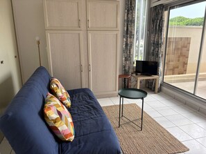 Ein Wohnzimmer mit einer Couch, einem Fernseher auf einem Schrank, einem gemusterten Teppich und Blick durch ein Fenster.