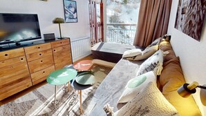 En moderne stue med et fladskærms-tv, træmøbler, en sofa med puder, et lille bord med en grøn underkop og et vindue med udsigt til træer og en balkon.