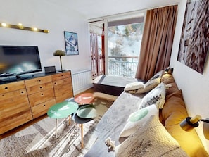A modern living room with a flat-screen TV, wooden furniture, a sofa with cushions, a small table with a green coaster, and a window with a view of trees and a balcony. - Ski-in/ski-out apartment, exceptional view - 6 guests (Les Arcs)