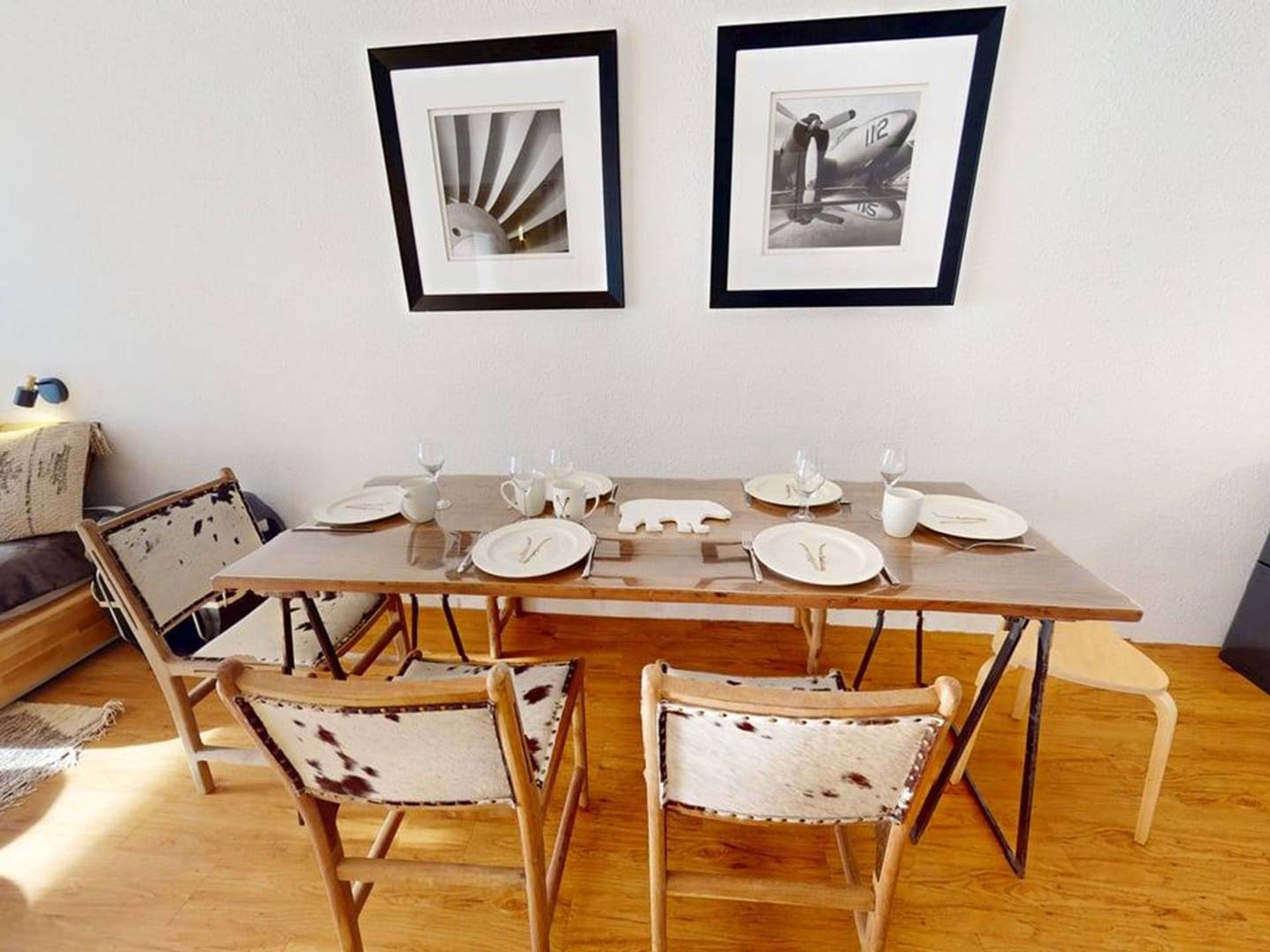 A dining area with a wooden table set for four, two framed pictures on the wall, and vintage-style chairs.