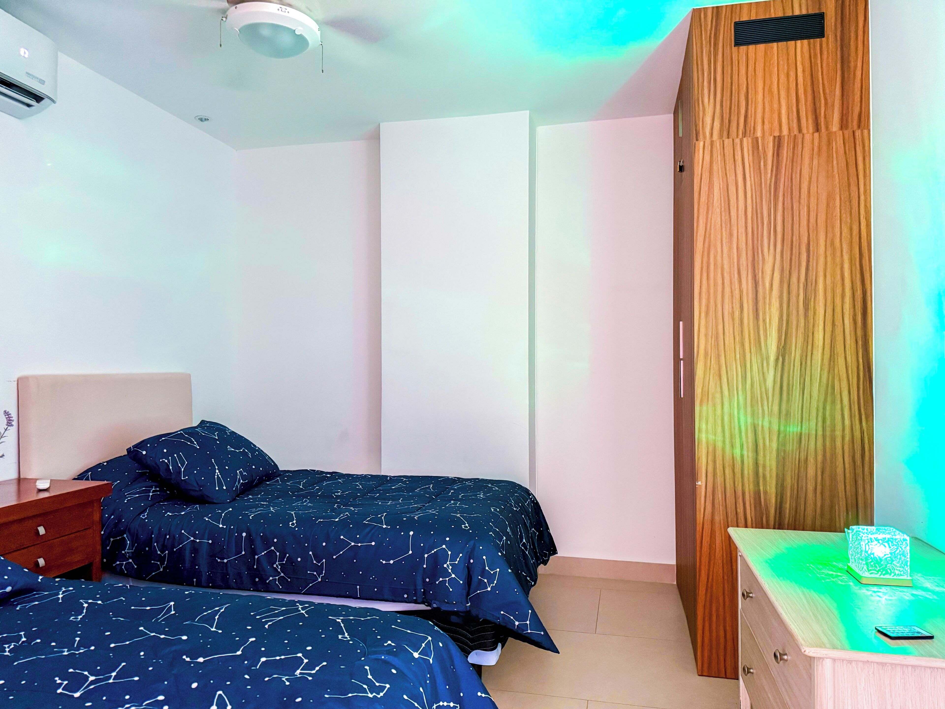 Quarto com duas camas, guarda-roupa de madeira, mesa de cabeceira e luminária no teto.