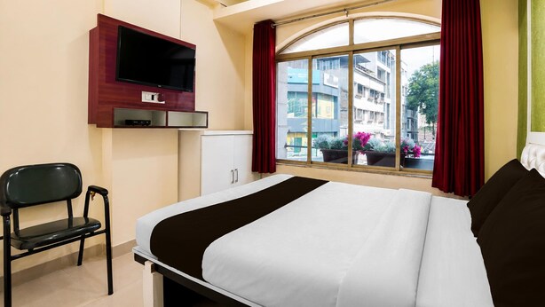 Habitación de hotel con cama, televisor montado en la pared, una silla, ventana con cortinas y vista a edificios.