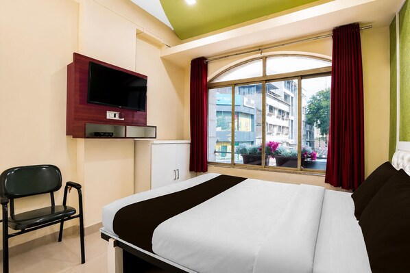 A hotel room with a bed, a TV mounted on the wall, a chair, a window with curtains, and a view of buildings outside.