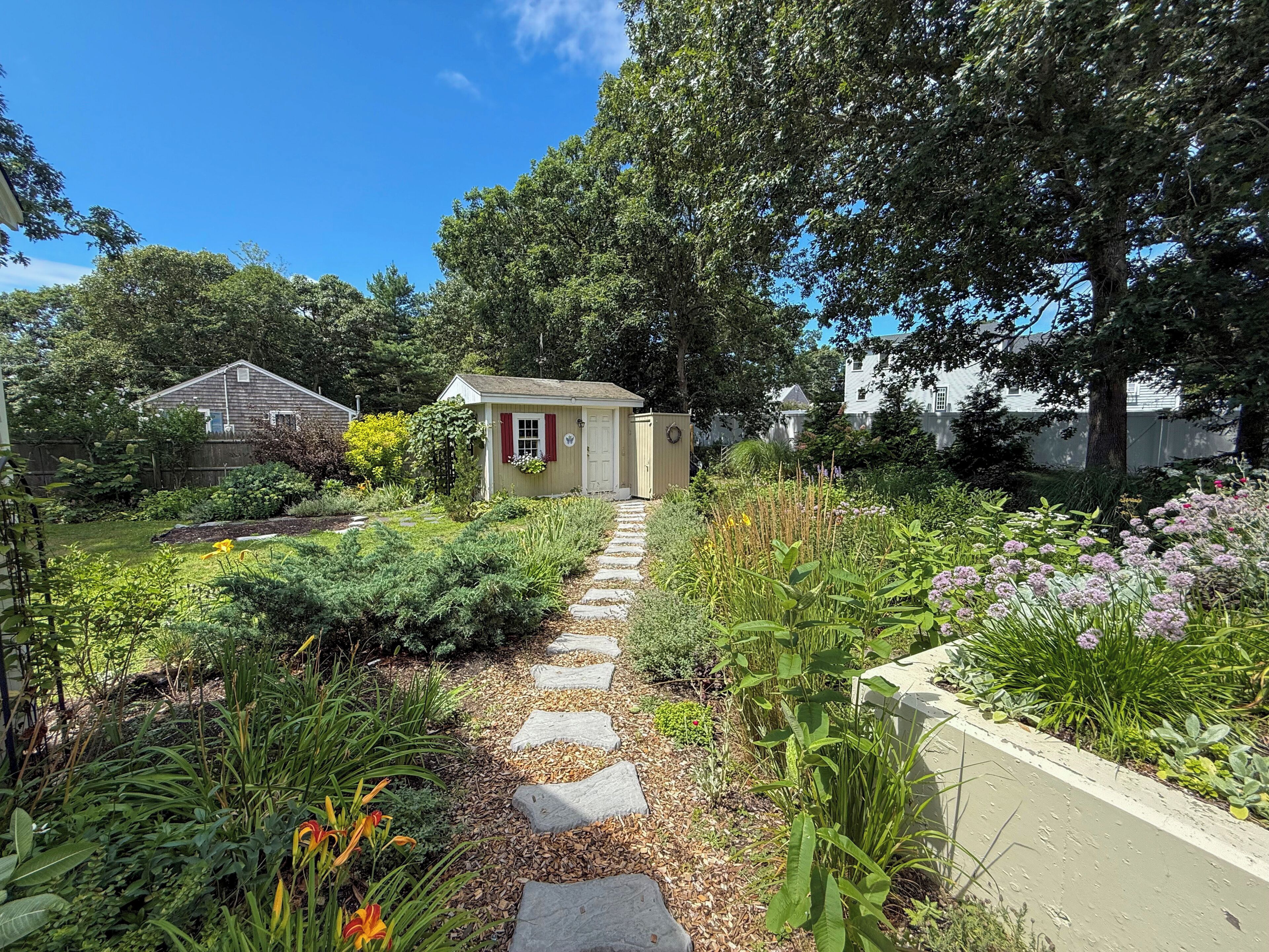 A garden pathway leading to a small shed with a door and windows, surrounded by various plants and a fence.
