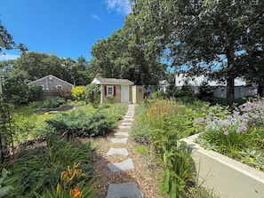A garden pathway leading to a small shed with a door and windows, surrounded by various plants and a fence. - Nautical three bedroom sleeping 6 in West Dennis! (West Dennis)