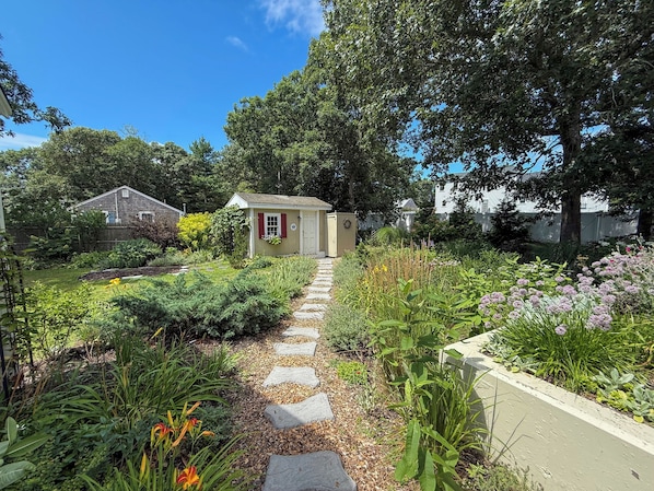 A garden pathway leading to a small shed with a door and windows, surrounded by various plants and a fence. - Nautical three bedroom sleeping 6 in West Dennis! (West Dennis)