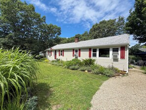 A single-story house with a white exterior and red shutters, surrounded by a well-maintained lawn and various plants. - Nautical three bedroom sleeping 6 in West Dennis! (West Dennis)