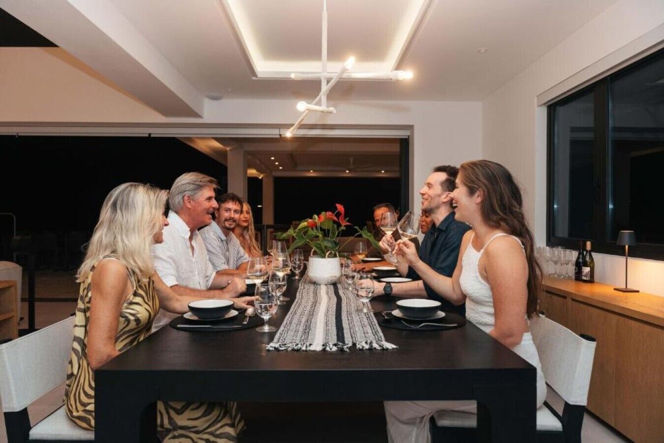 Een groep mensen dineert aan een tafel met een modern ontwerp, met een centrale vaas vol bloemen, een lange tafel en een moderne plafondlamp.