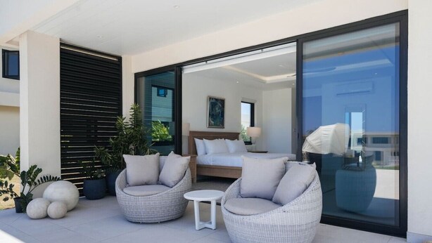 Un espace extérieur moderne aménagé avec des chaises en osier et une petite table, une chambre dotée d’un lit et d’une table de chevet, et une grande porte-fenêtre coulissante donnant sur un balcon agrémenté de plantes en pot.