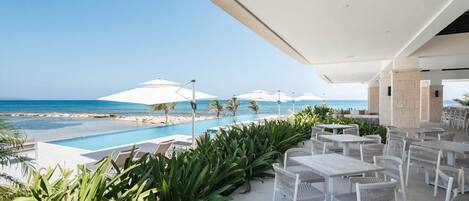 Un área de estar al aire libre con mesas y sillas, piscina y sombrillas con vista al océano.