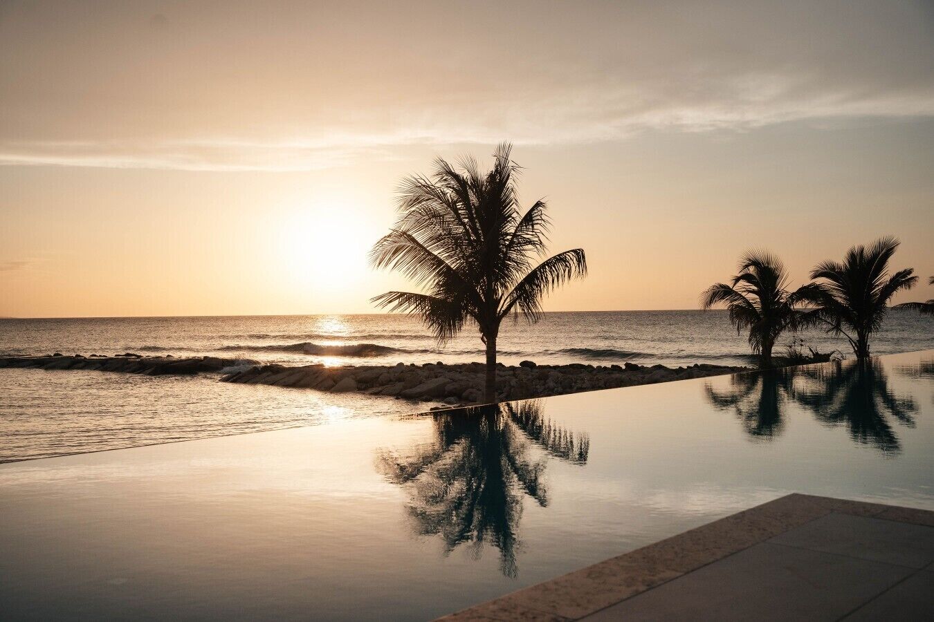 A serene beachfront scene with a reflective pool, palm trees, and a calm ocean at sunset.