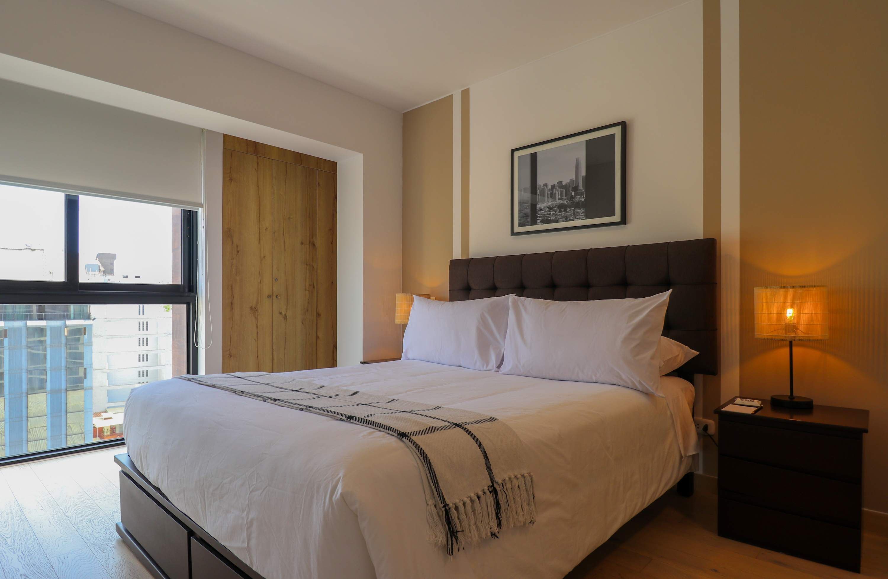 A hotel room with a large bed, a nightstand with a lamp, a framed cityscape picture on the wall, and a view of buildings through the window.