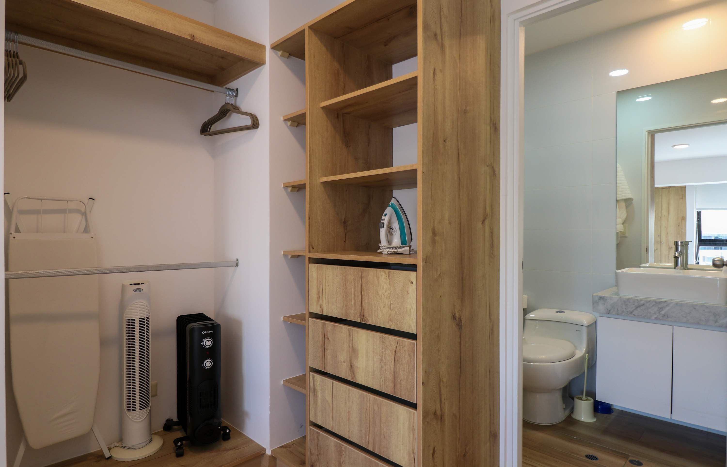 Baño con inodoro, lavamanos y espejo. Se observa una estantería de madera con un perchero y un ventilador.