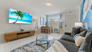 Una sala de estar con televisión mostrando una escena de playa, una mesa de centro de vidrio, un sofá y un comedor con sillas y una mesa.