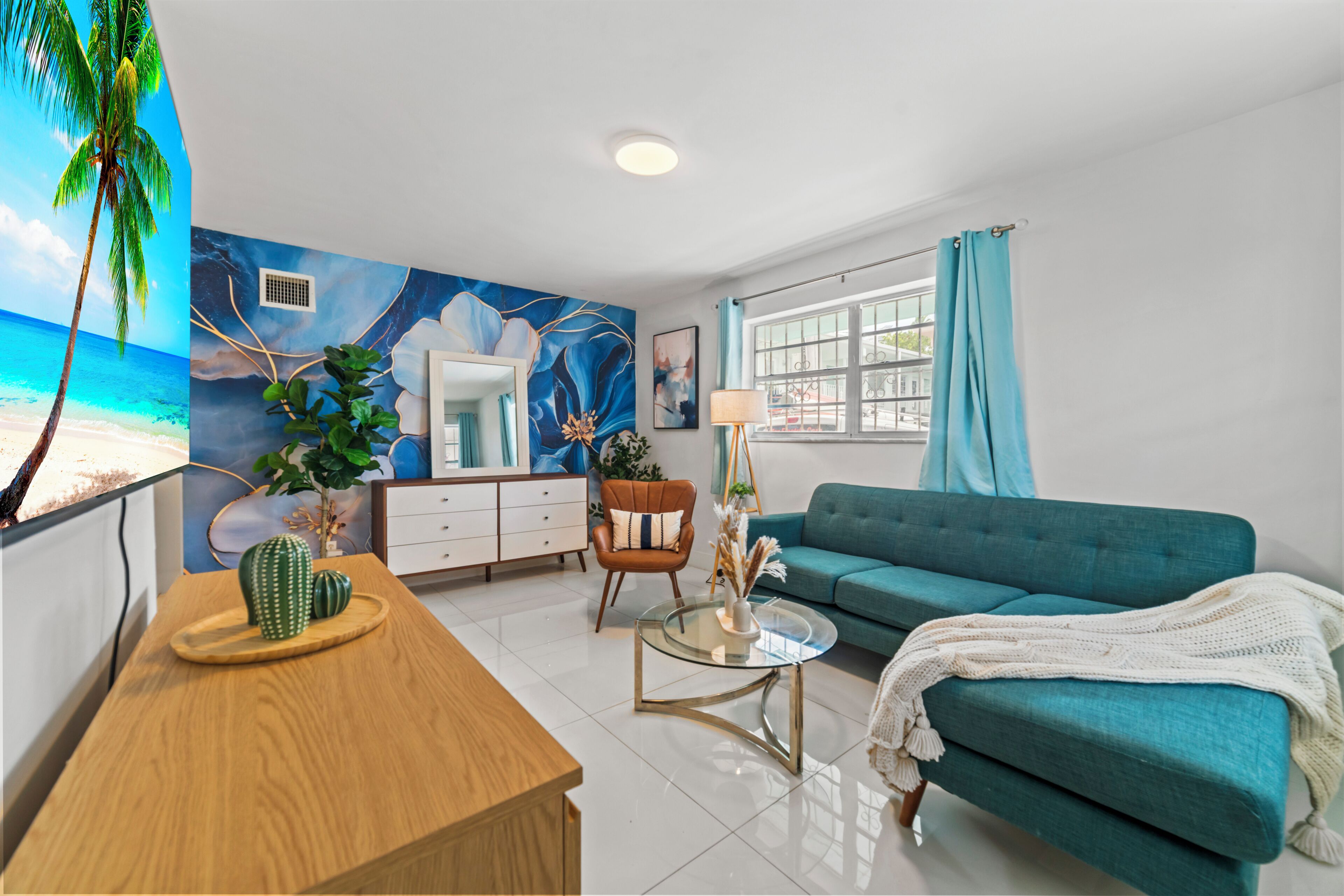 Uma sala de estar moderna com um sofá verde-água, uma mesa de centro de madeira, uma mesa de apoio com tampo de vidro, um espelho e uma parede com um mural tropical.