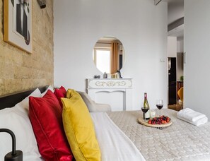 Ein Hotelzimmer mit Bett, einem runden Spiegel, einer weißen Kommode und einem Frühstückswagen mit Obst und Wein.