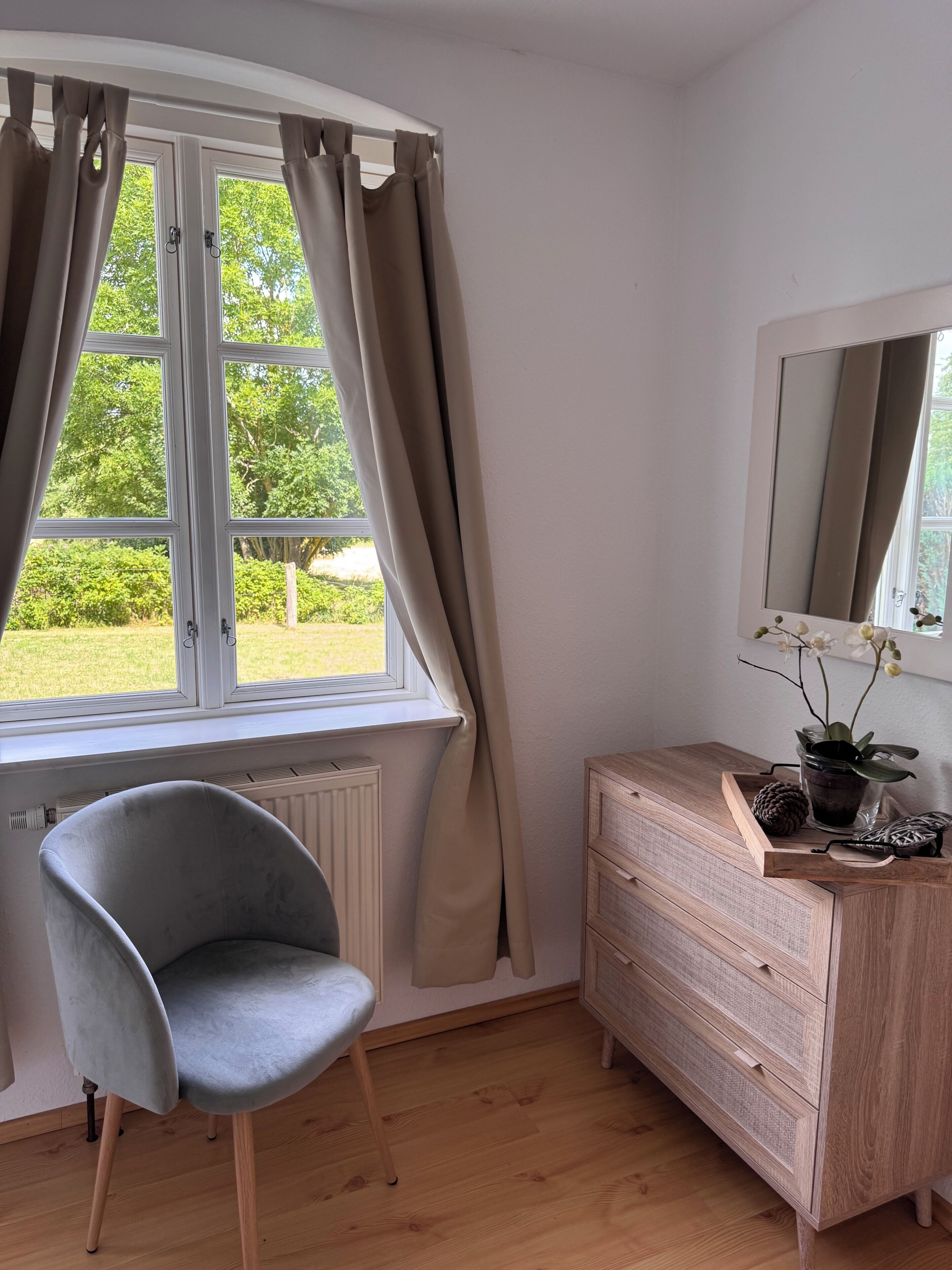 Ein Zimmer mit Fenster, Stuhl, Kommode und Spiegel.