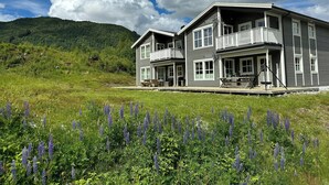 Una casa a due piani con un balcone, circondata da un campo di fiori viola e una collina boscosa sullo sfondo.