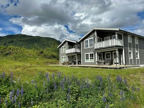 Ein zweistöckiges Haus mit Balkon, umgeben von einem Feld purpurner Blumen, mit einem bewaldeten Hügel im Hintergrund.