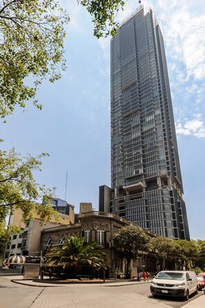 Un grand immeuble moderne à la façade de verre, situé à côté d’un bâtiment historique à la façade classique, avec un palmier devant.