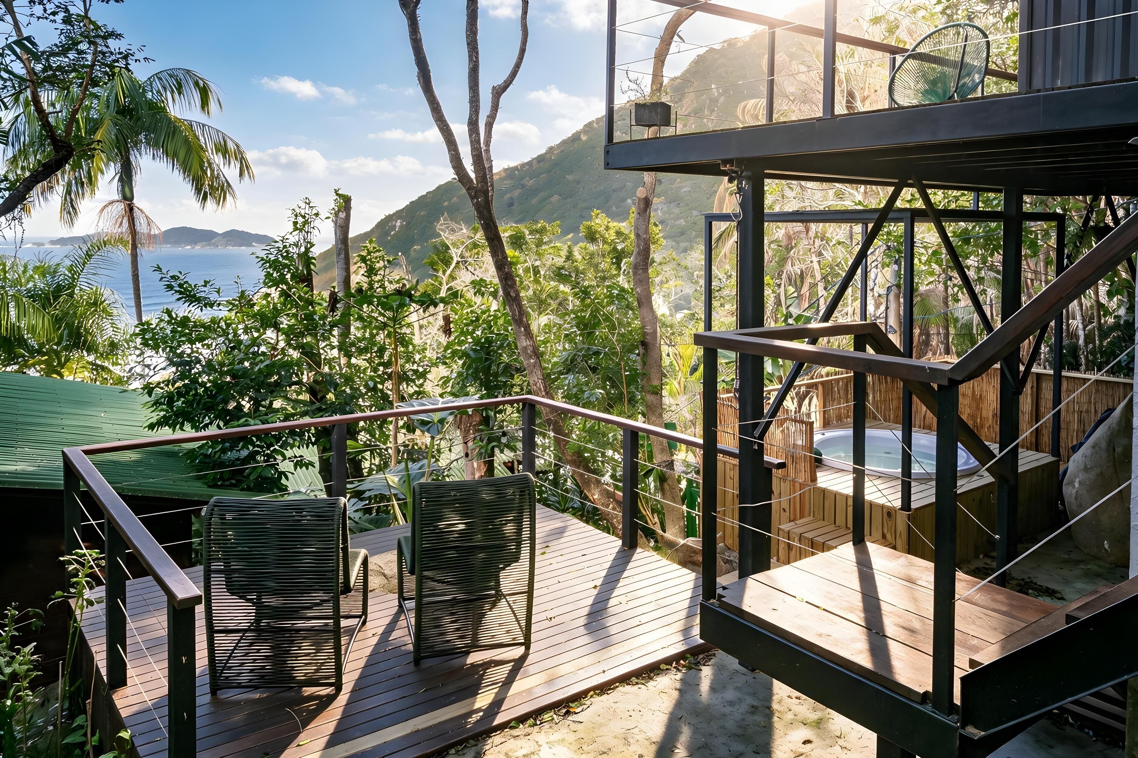 A wooden deck with two chairs, a hot tub, and a view of the ocean and hills.