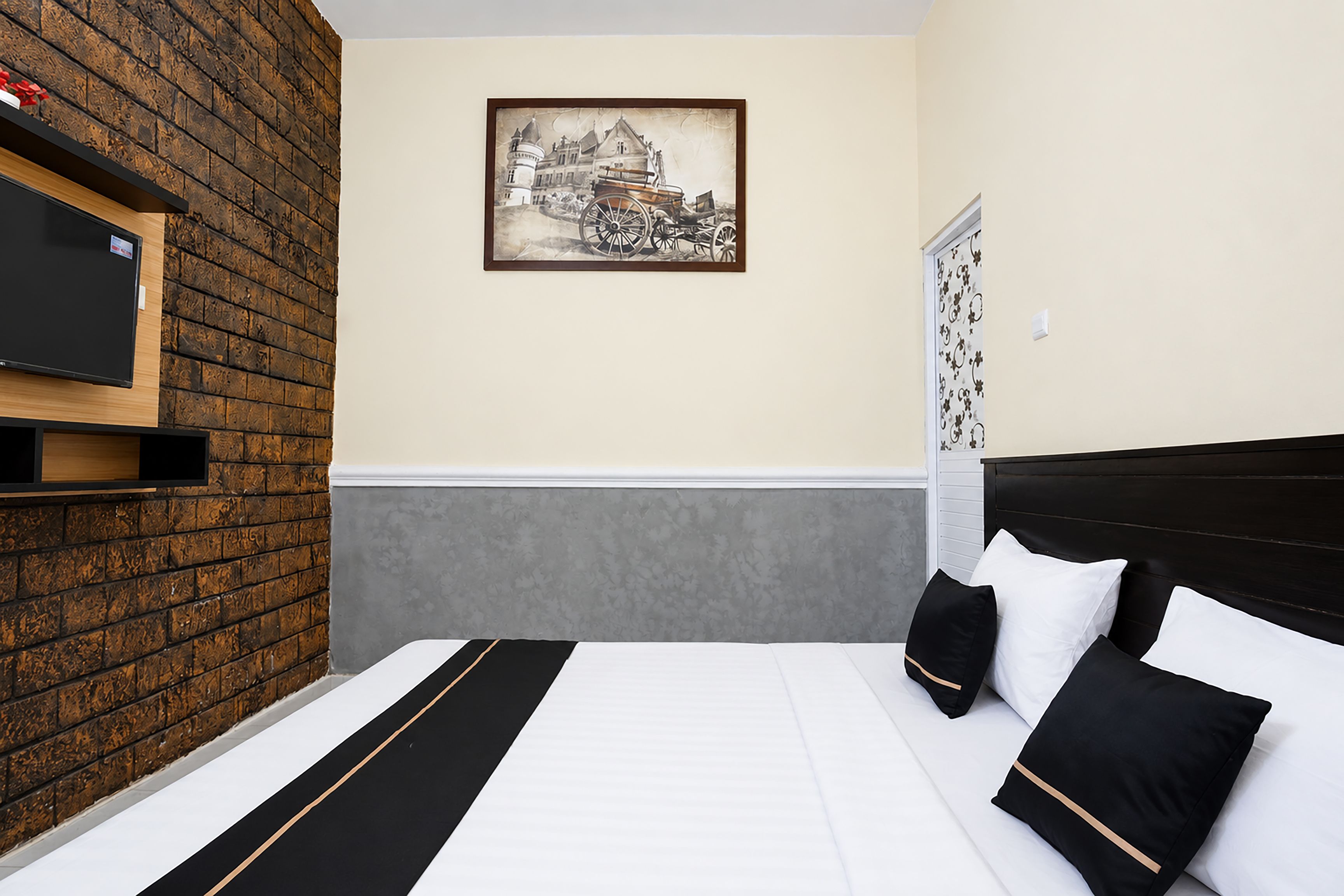 A hotel room with a brick wall, a framed picture, a bed with black and white bedding, and a TV mounted on the wall.