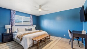 Ein Schlafzimmer mit Bett, Nachttischen, Schreibtisch, Stuhl, Deckenventilator und einem Fenster mit Vorhängen.