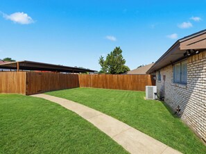 Un jardin avec une clôture en bois, un chemin en béton et un bâtiment en briques.