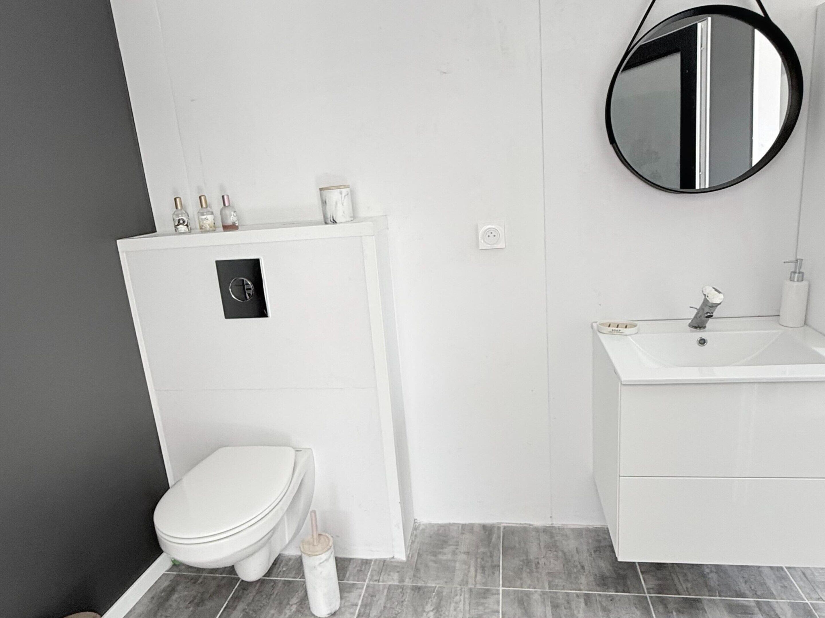 Une salle de bain équipée d’un WC, d’un lavabo, d’un miroir et d’une petite étagère où sont rangés les articles de toilette.
