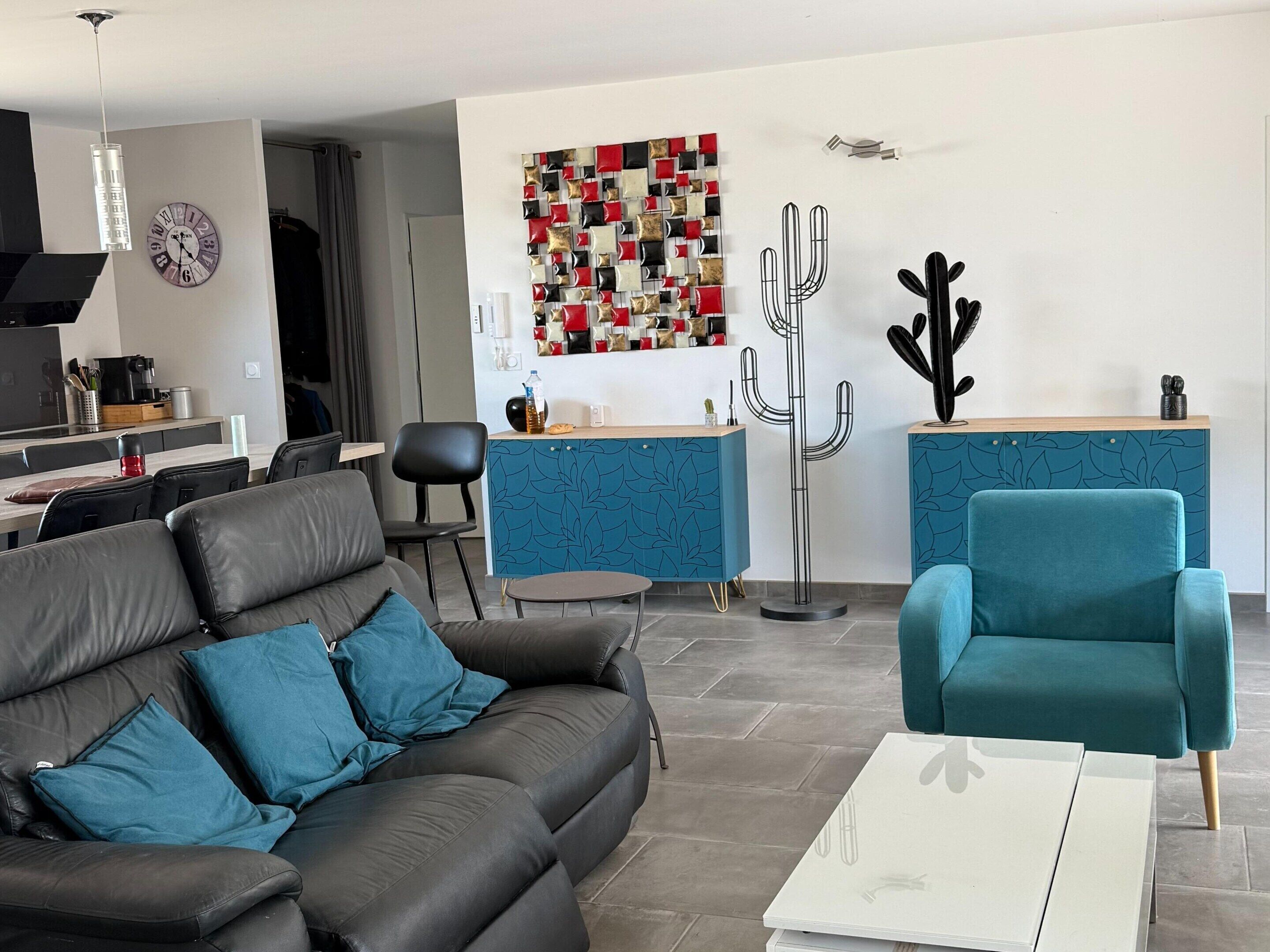 Een moderne woonkamer met een bank, een salontafel, een blauwe fauteuil en een keukenhoek met een blauwe kast.