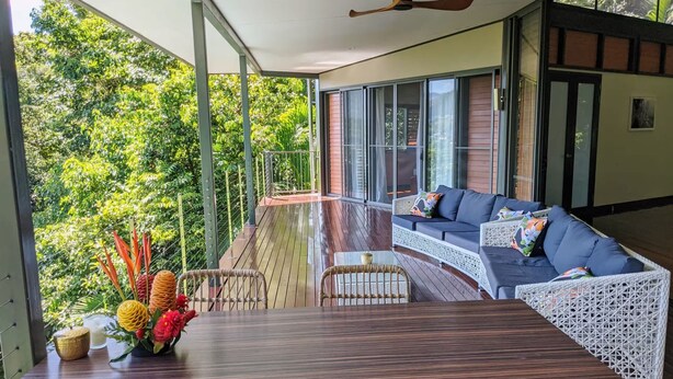 A covered outdoor seating area with a wooden table and chairs, a sofa with cushions, and a view of lush greenery. - Rainforest Meets Luxury - Infinity Pool Views (Cairns)