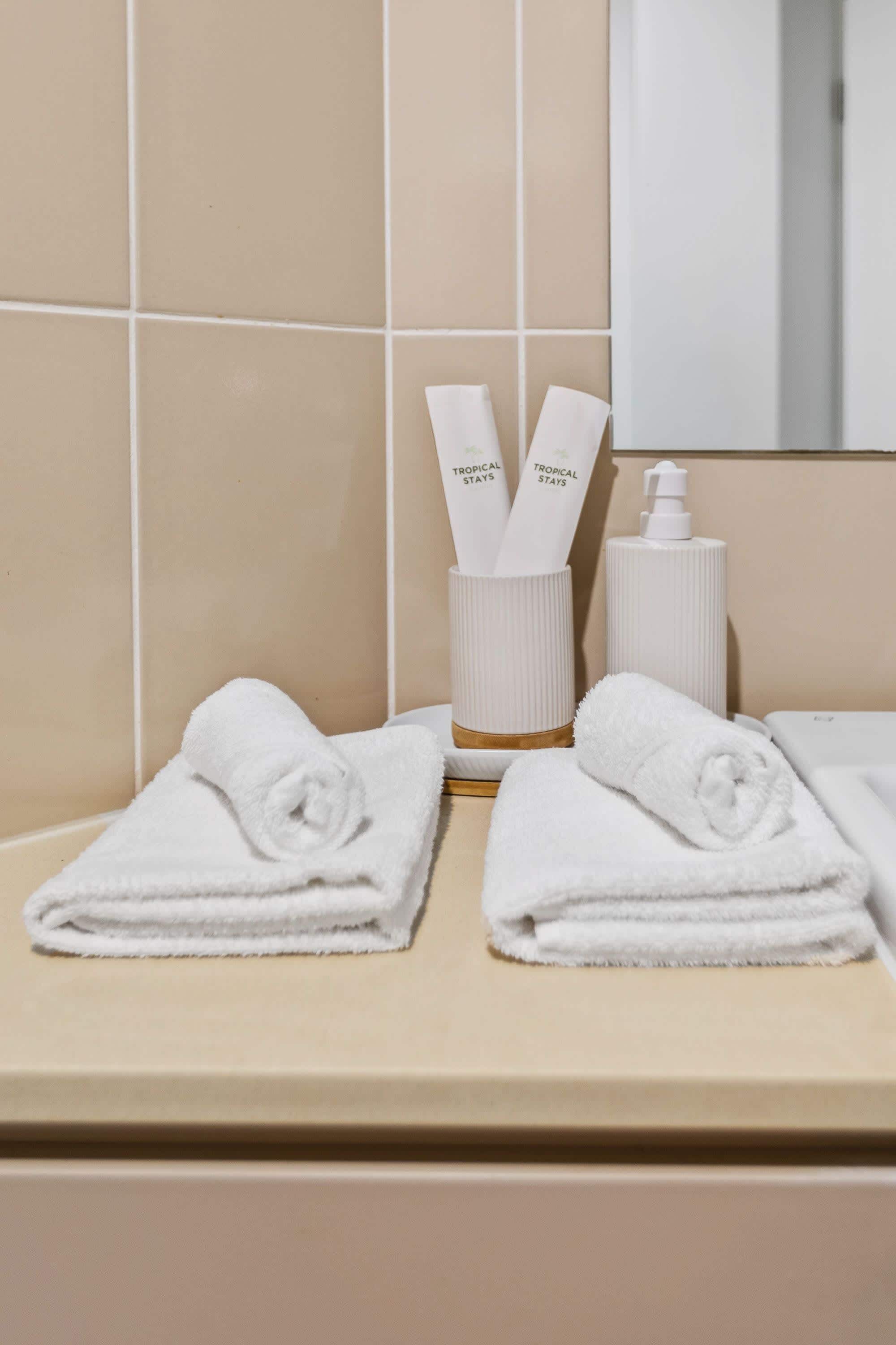 En badeværelsesbordplade med to pænt foldede hvide håndklæder, en sæbe og en lotion.