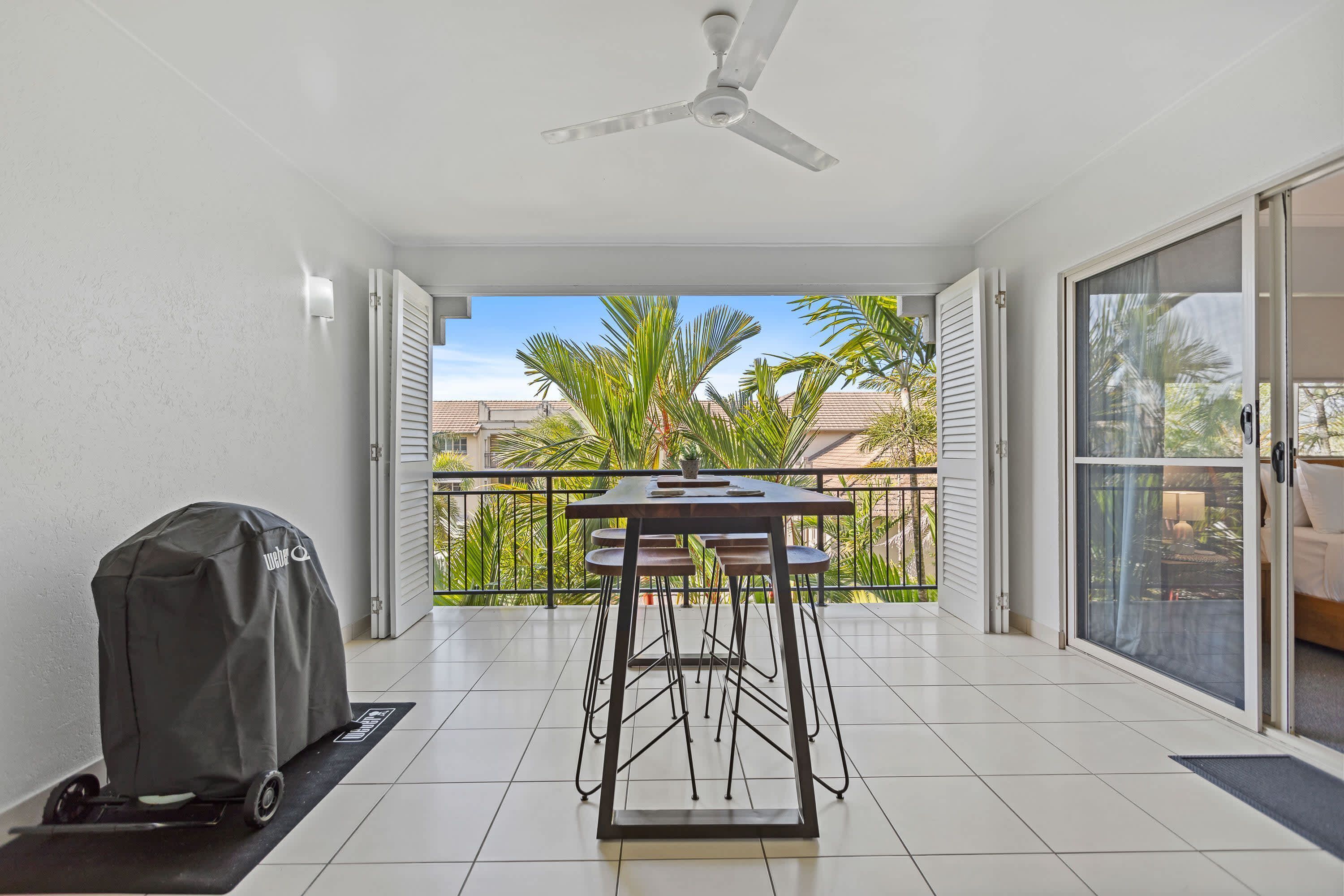 A balcony with a table and chairs, a grill, and a view of palm trees and buildings.