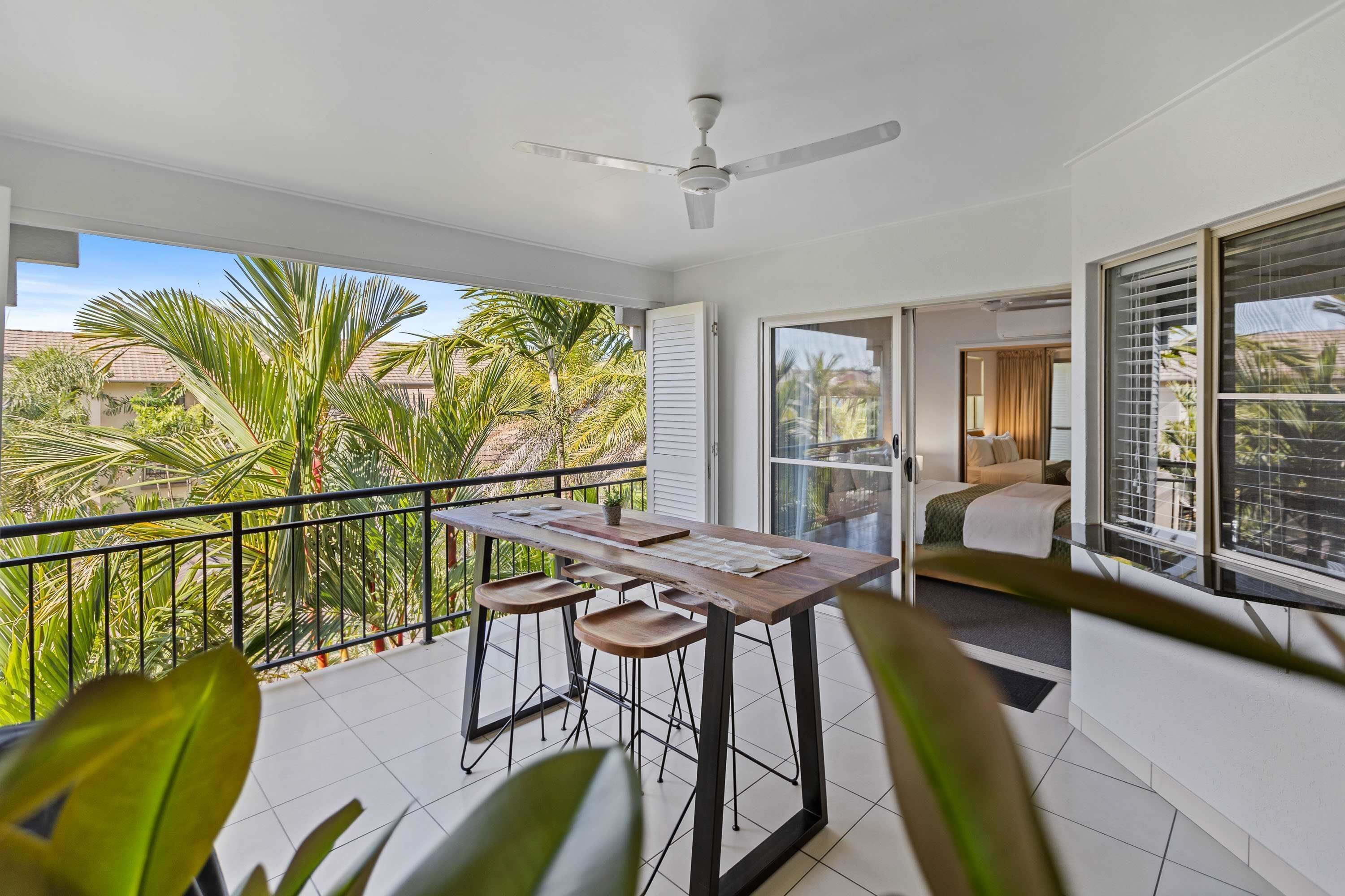 A balcony with a table and chairs, a ceiling fan, and a view of palm trees and houses.