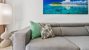 Ett rum med en grå soffa, en grön kudde, en mönstrad kudde, en vit lampa och en tavla med ett tropiskt ölandskap som hänger på väggen.