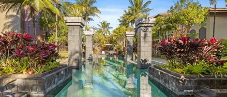 A swimming pool with clear water, surrounded by tropical plants and palm trees, with a view of a building in the background.