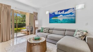 Ett modernt vardagsrum med en grå soffa, ett runt soffbord i trä och en stor tavla med ett tropiskt strandmotiv på väggen.