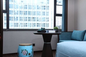 A room with a view of a building through large windows, a blue drum, a small table with a plant, and a blue sofa.