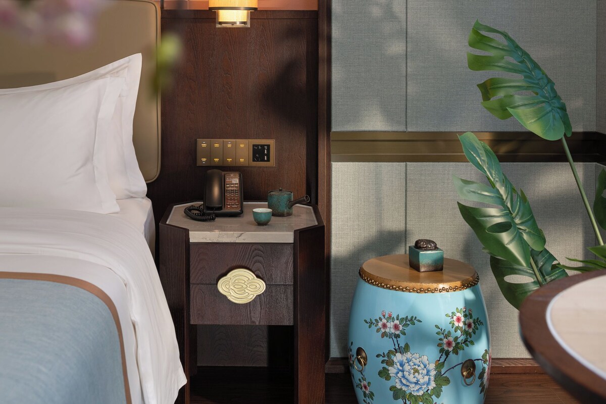 Una habitación de hotel con cama, mesita de noche con lámpara, teléfono y una planta decorativa.