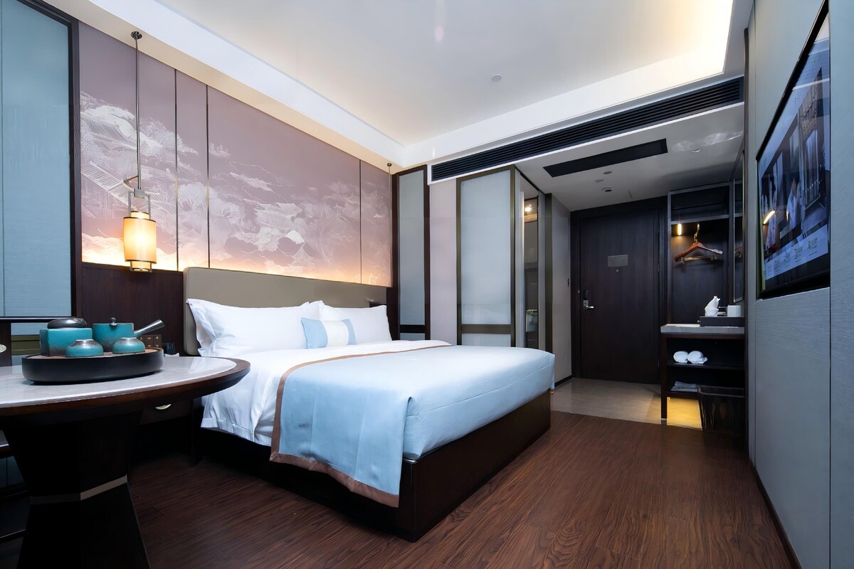 Una habitación de hotel moderna con una cama grande, una mesita de noche con lámpara, una obra de arte en la pared y vistas al pasillo.