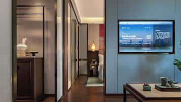 Un pasillo de hotel con una pantalla de televisión mostrando información, una cama, una mesita de noche y una pequeña mesa con objetos decorativos.