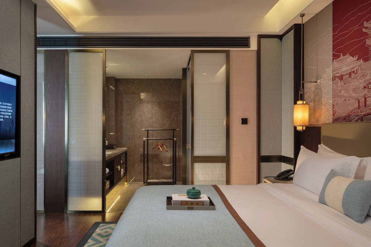 Una habitación de hotel con cama, mesita de noche, lámpara y vista al baño.