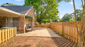 A backyard with a patio, a brick building, a wooden fence, and a large tree.