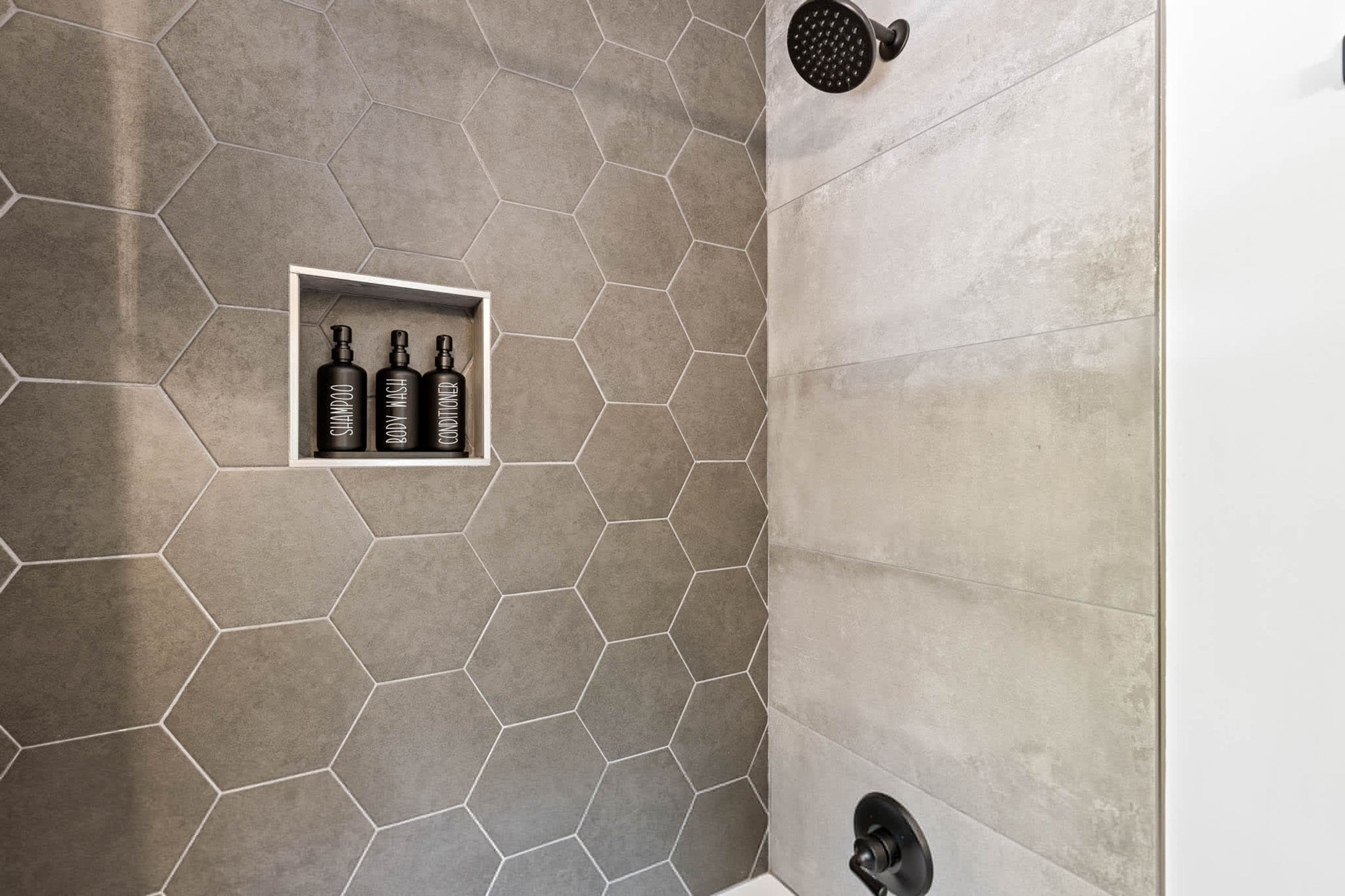 Un baño con azulejos hexagonales, una ducha y un estante con tres botellas de champú, gel de baño y acondicionador.
