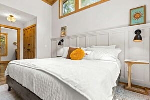 A bedroom with a large bed, a wooden headboard, a nightstand, a wall-mounted lamp, and a framed picture.