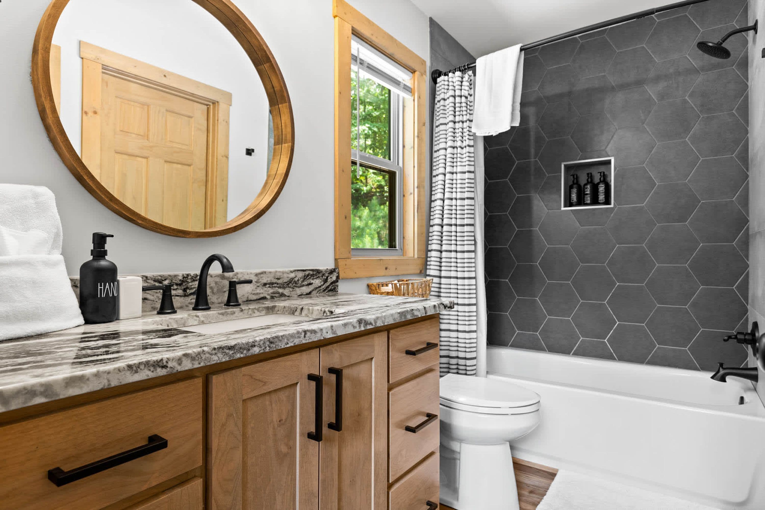 Un baño con un tocador de madera, un espejo redondo, encimera de mármol, inodoro y ducha con pared de azulejos hexagonales.