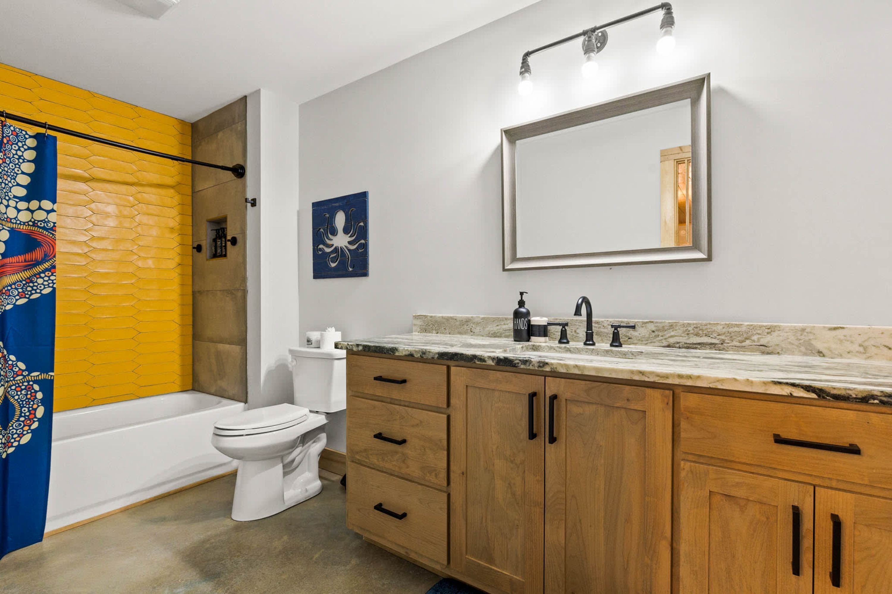 Un baño con un inodoro blanco, un tocador de madera con encimera de mármol, un espejo y una ducha con pared amarilla.