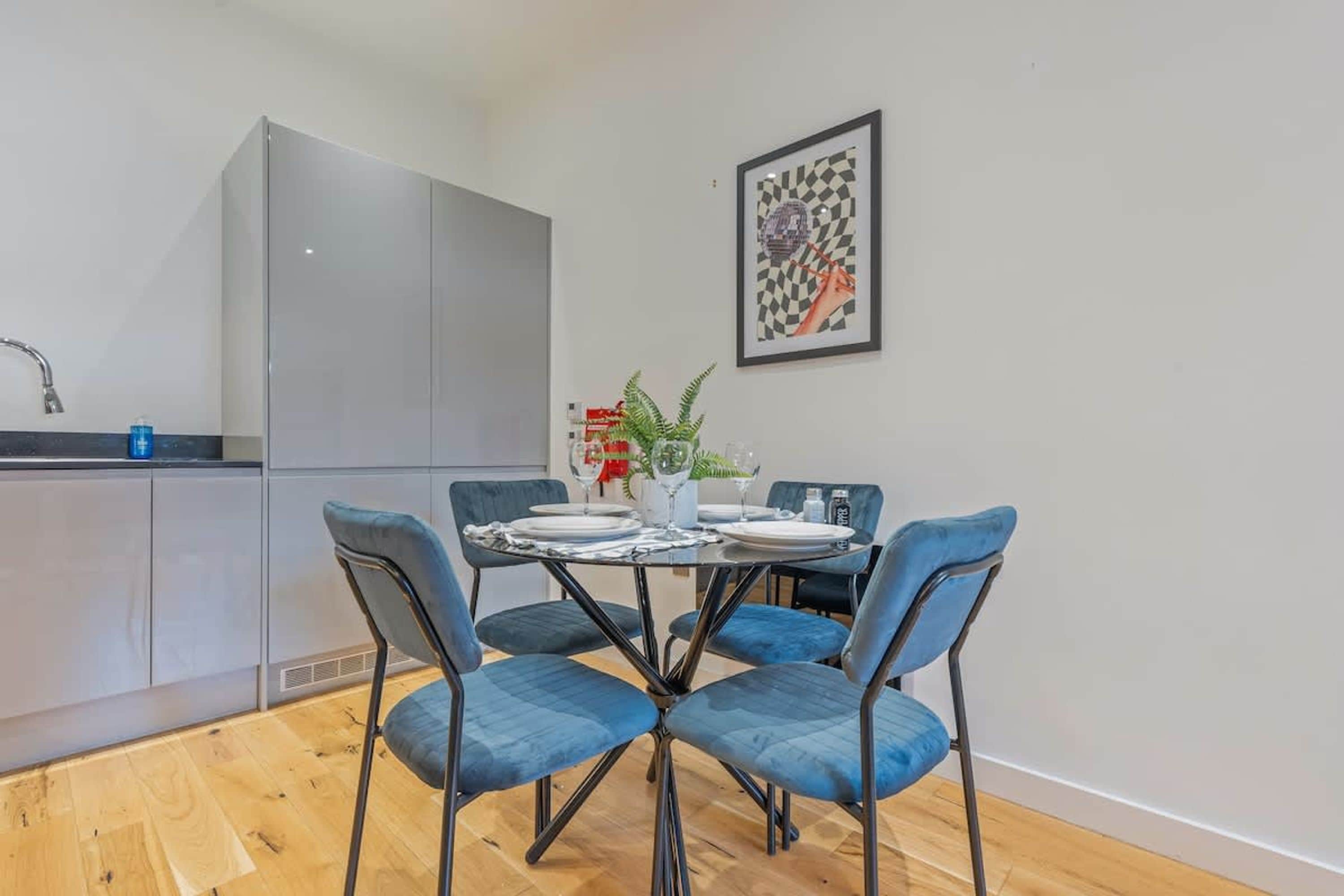 A compact kitchen with a dining area, featuring a table set for two with blue chairs, a potted plant, and a framed artwork on the wall.